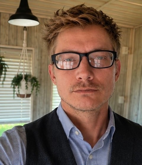 Man wearing glasses and dark cardigan over light blue shirt, indoors with pendant lamp and hanging plant visible in background